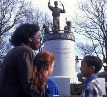 A Black woman, a white girl, and a Black boy in the foreground with the Arthur Ashe Monument in the background.