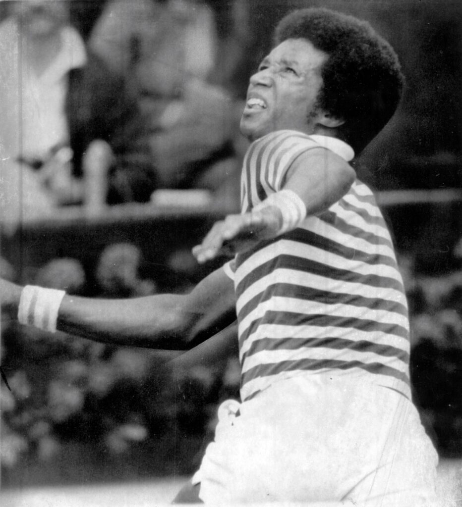 Arthur Ashe going for a forehand shot at the US Open. He is wearing a striped polo shirt and white shorts.