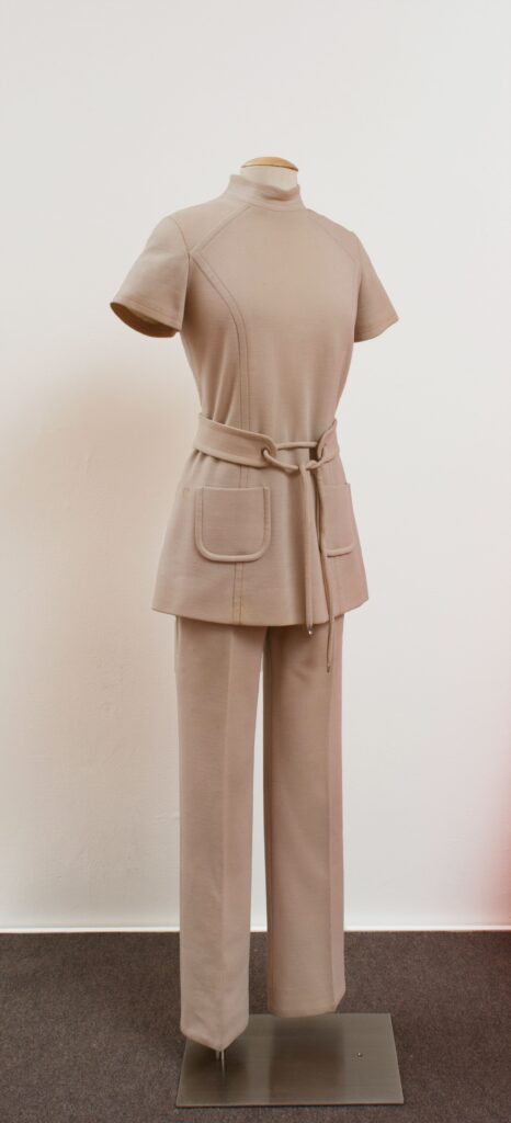 Short-sleeved, mock turtleneck, cream-colored pantsuit with a tie belt and two pockets on the front of the shirt.