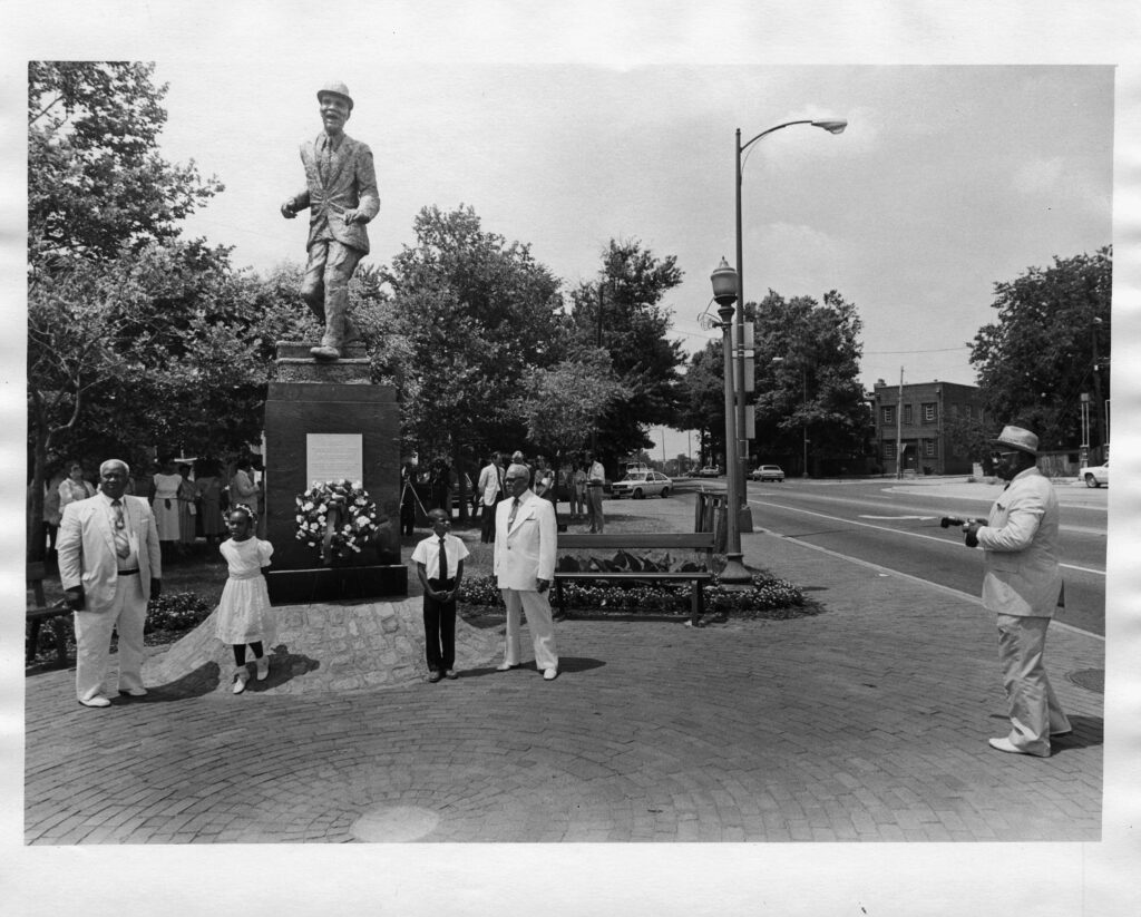 Three men in white suits and two children at the dedication of the Bill "Bojangles" Robinson statue which is in the background. The statue is of a man wearing a bowler hat dancing. 