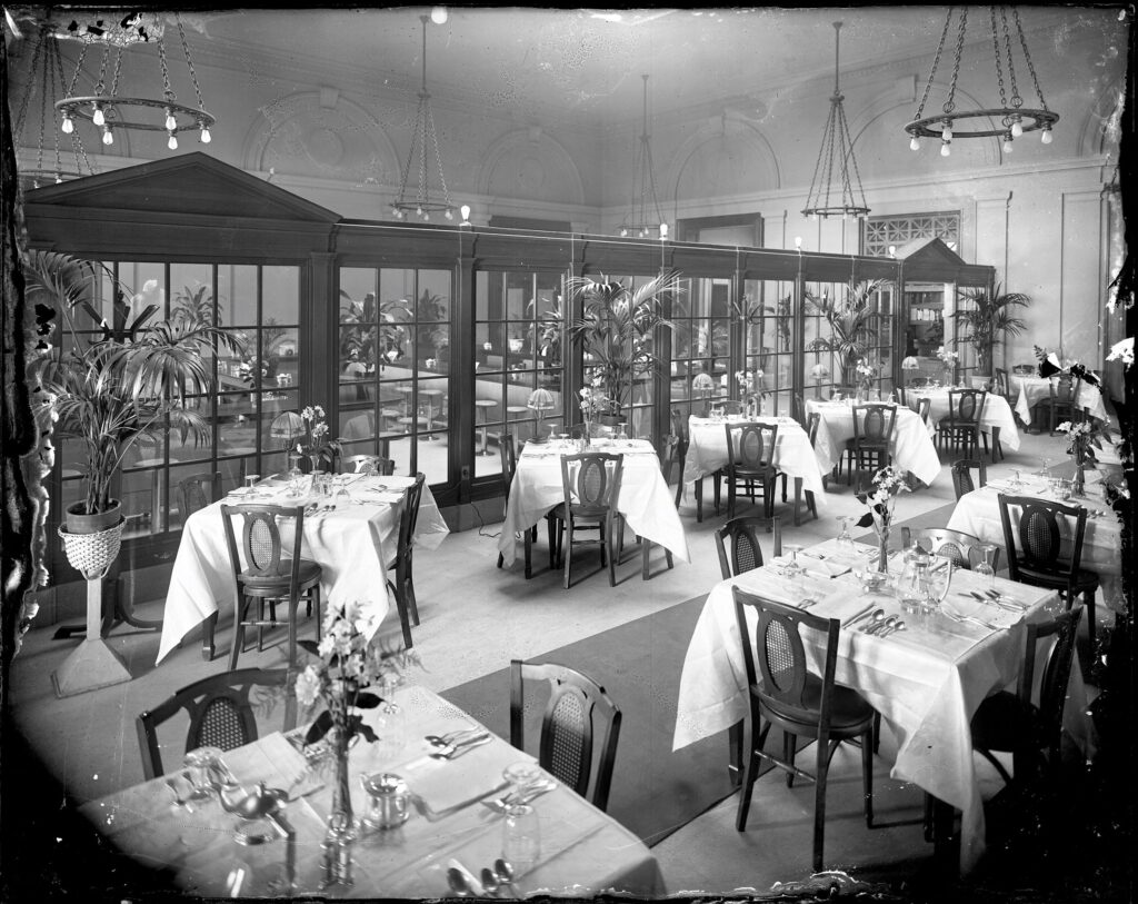 Dining room with tables for four set with tablecloths, silverware and glasses. A mirrored window wall runs the length of the room. There are four tall potted plants arranged along the wall.