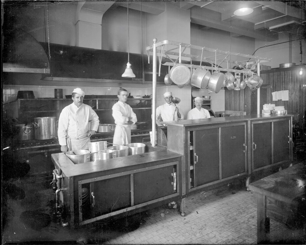 Four men in white chef's coats standing in a kitchen with multiple pots on the stove and many others hanging from hooks.