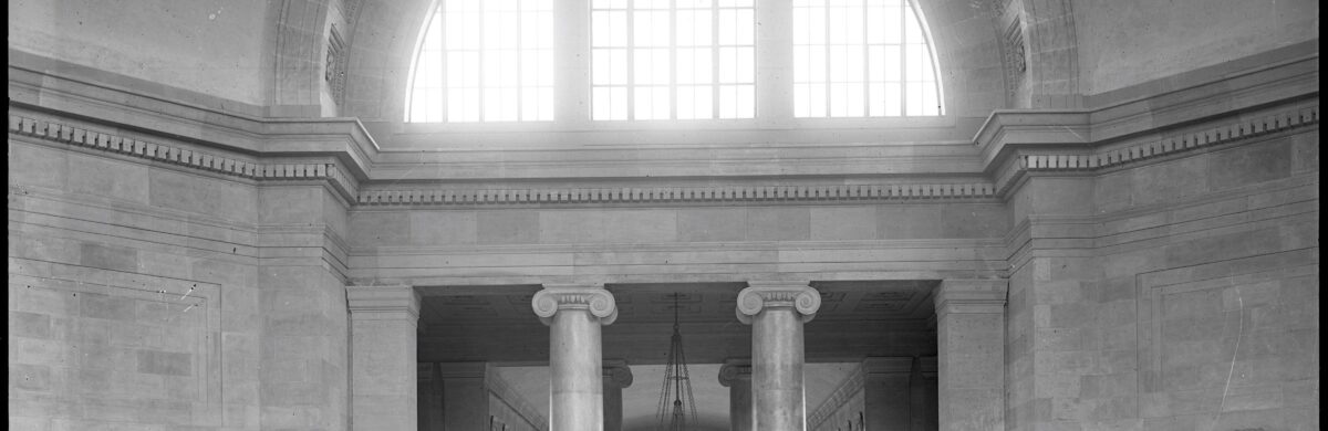 Waiting room in Broad Street Train Station. Two large ionic columns and four long wooden benches. A globe hangs above and the ceiling is tall.