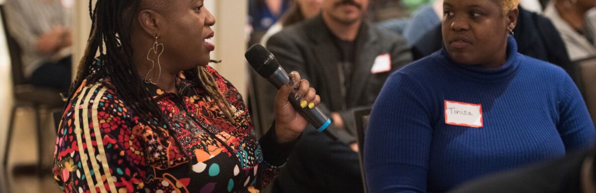 An audience of an event is depicted. In the foreground, a Black woman audience member holds the microphone and speaks. Next to her another audience member listens.