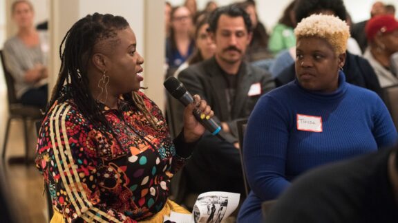 An audience of an event is depicted. In the foreground, a Black woman audience member holds the microphone and speaks. Next to her another audience member listens.