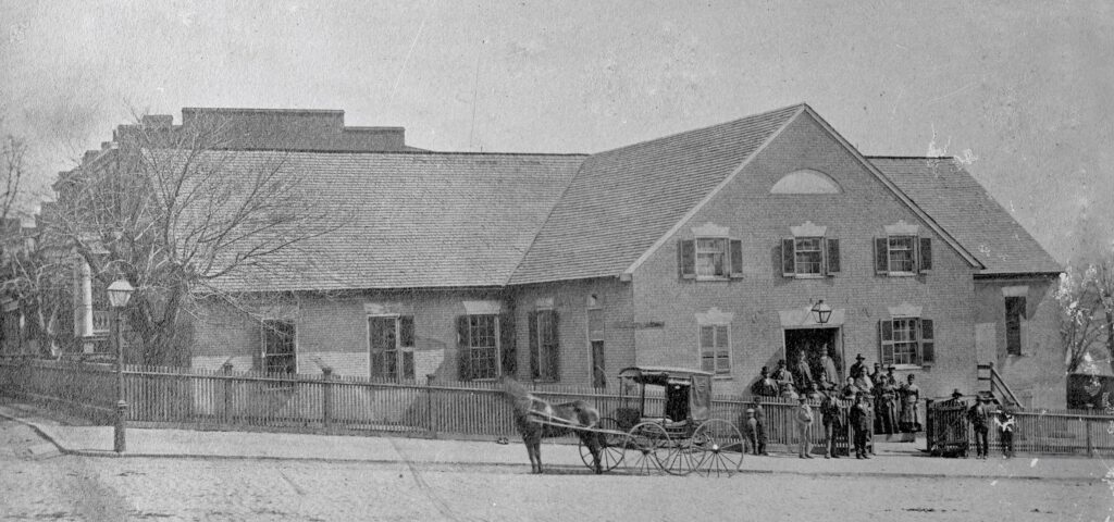 The church building is brick and predominately one-story with the front portion of the church being two- stories. There are black shutters on most of the windows. A group of 15-20 men, women and children stand outsid the front door and fence. A horse drawn carriage is to the left of the door.