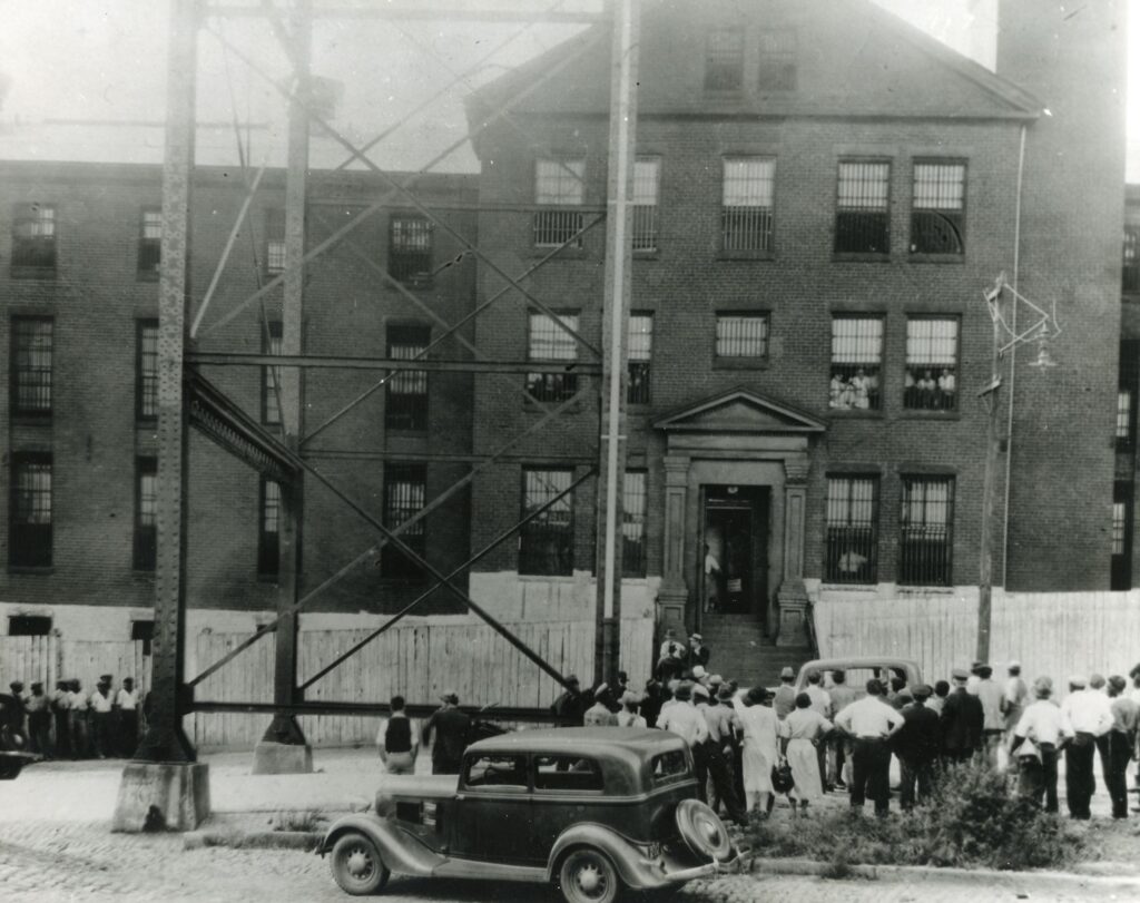 The jail is a three-story large brick building with a crowd of people out front facing the jail. there is one car in the foreground. 