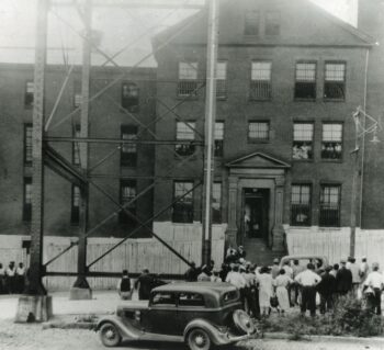 The jail is a three-story large brick building with a crowd of people out front facing the jail. there is one car in the foreground. There are bars on all the windows.