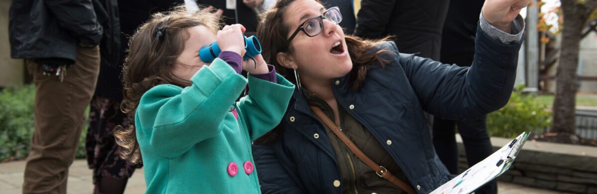 A young child is looking through binoculars at something unidentified while their parent points excitedly. There is a tour group standing in the background.