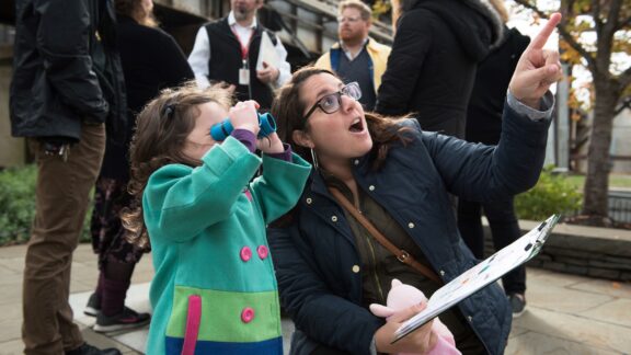 A young child is looking through binoculars at something unidentified while their parent points excitedly. There is a tour group standing in the background.