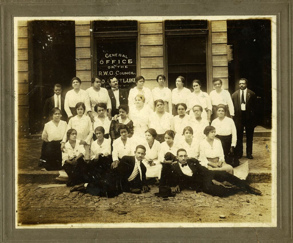 Staff photo in front of the St. Luke's General Office. Two men are in front with three rows of women with Maggie L. Walker in the middle. 