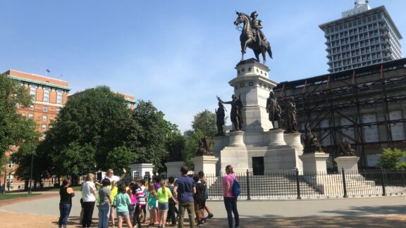 A group of children and adults is shown gathered in a circle and conversing. Behind them is the monument of George Washington on horseback at the Virginia State Capitol.