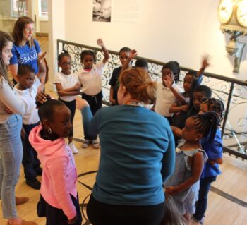 A group of students and teachers are gathered in a circle in the museum. Young students are raising their hands while the adults point at them.