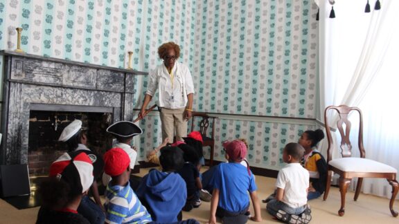 Young students sit on the floor in a historic room with their backs to the camera. In front of them a Valentine Educator stands showing the students some historic objects.