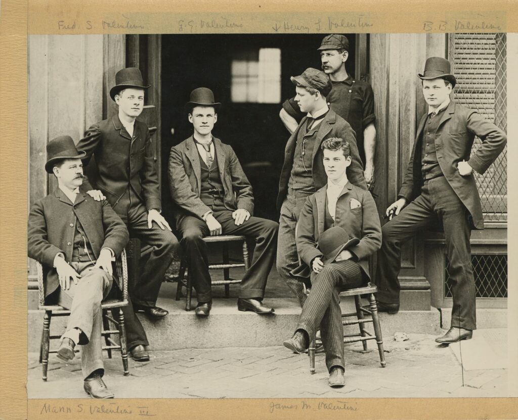 7 Valentine Brothers either sitting in chairs or standing outside of the factory. Many are dressed in suits and wearing hats. 