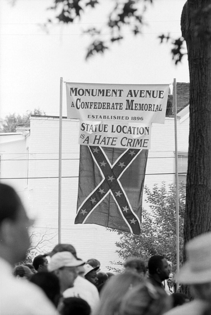 People in the foreground and a Confederate battle flag and sign in the background that reads: “Monument Avenue a Confederate Memorial established 1896 Statute Location is a Hate Crime.”