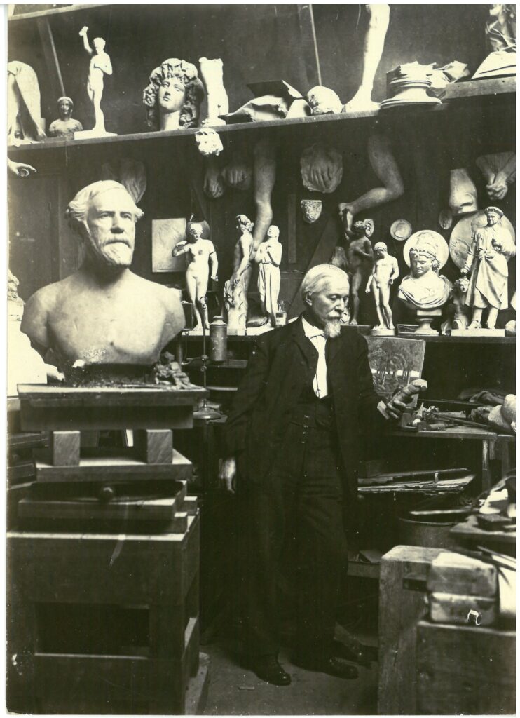 As an older gentleman, Edward Valentine stands in his studio dressed in a a black suit with white shirt surrounded by his sculpture including a large bus of Robert E. Lee.