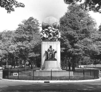 The Maury Monument with a bronze globe on top of a pedestal with a figure of a seated Maury in the fenced median of Monument Avenue with trees.