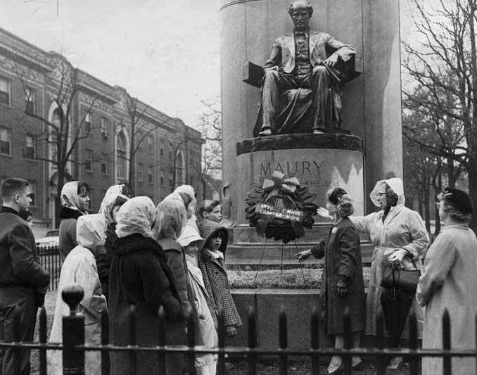 Women and children standing in the rain in front of the Maury Monument with a wreath.