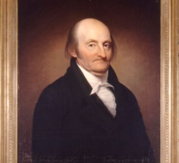 Portrait of Dr. James McClurg with a golden frame. He is wearing a black coat with a high collared shirt tied around his neck. He is predmoniately bald with deep set eyes.