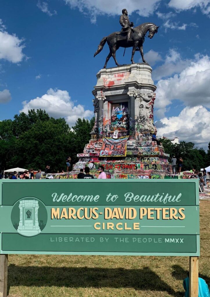 A green sign for Marcus-David Peters Circle in front of the graffiti-covered Lee Monument.