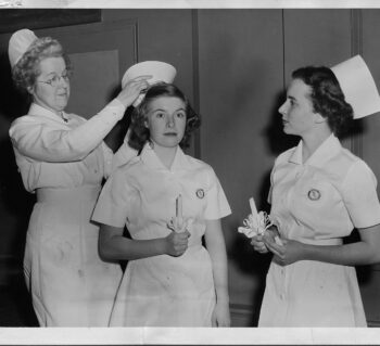 One nurse is putting on a cap for another nurse who is holding a candle. Another nurse holding a candle is watching. All are wearing white nurses uniforms.
