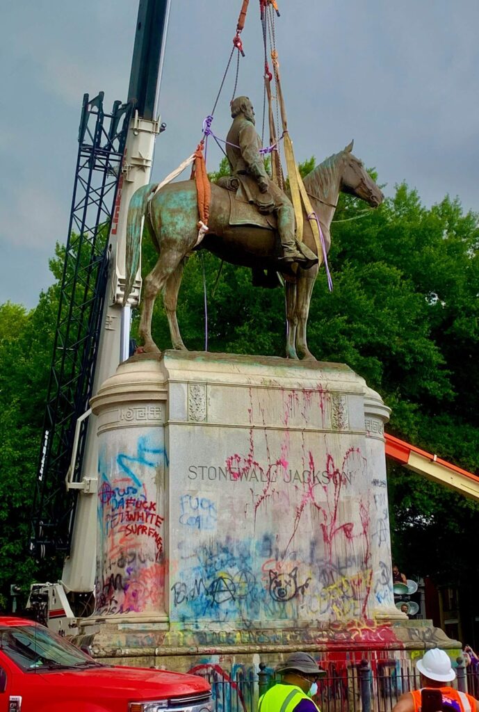 A crane in the background and workers in the foreground flank the graffiti-covered equestrian Stonewall Jackson Monument During a downpour, a clap of thunder rang out at the moment the crane lifted the statue from the pedestal followed by First Baptist Church ringing their bells. Team Henry removed the pedestal in early 2022 and paved over the intersection.