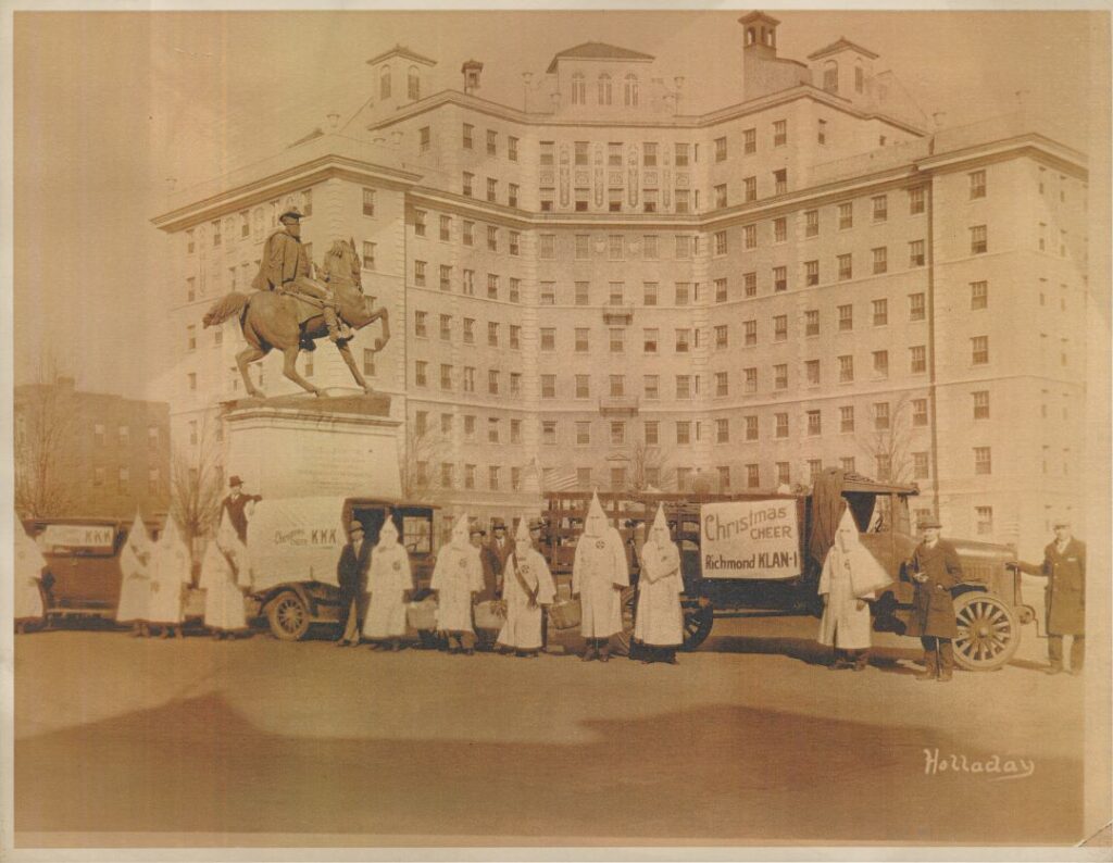 Men in white hoods and robes from the KKK stand in front of the Stuart Monument with men in suits beside trucks with posters on them reading “Christmas Cheer Richmond Klan 1.”