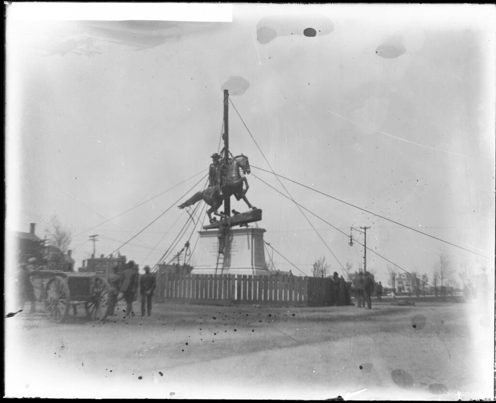 Men guiding a bronze statue of J.E.B. Stuart on a horse into place in Richmond while the statue hangs from ropes and pulleys on a tall derrick.