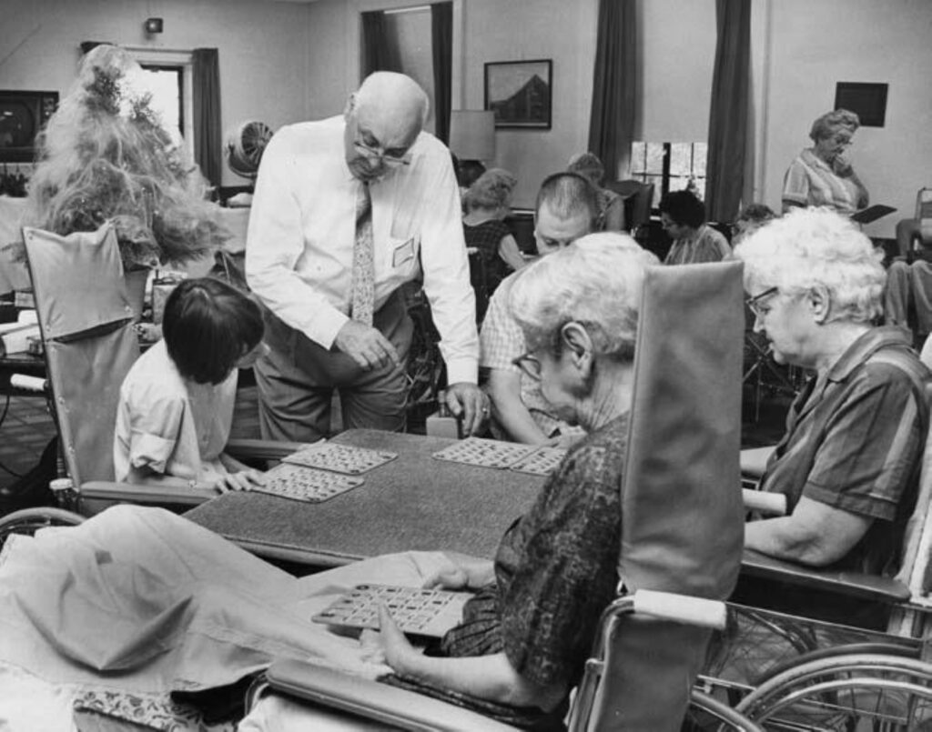 Older gentleman standing and helping a young woman, a man and two older women in wheelchairs play bingo.