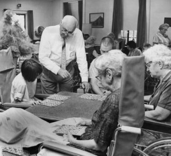Older gentleman standing and helping a young woman, a man and two older women in wheelchairs play bingo.