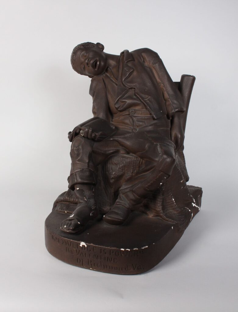Plaster sculpture painted brown depicting a Black boy asleep in a chair with an open book on his knee.