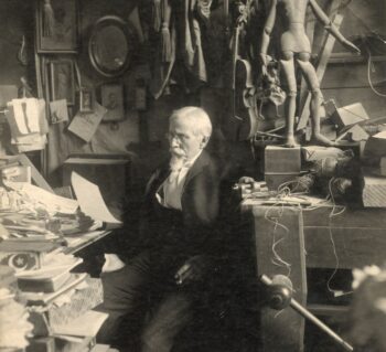 Edward Valentine reading in his studio surrounded by tools, papers, and a workbench with an artist’s mannequin sitting on top.