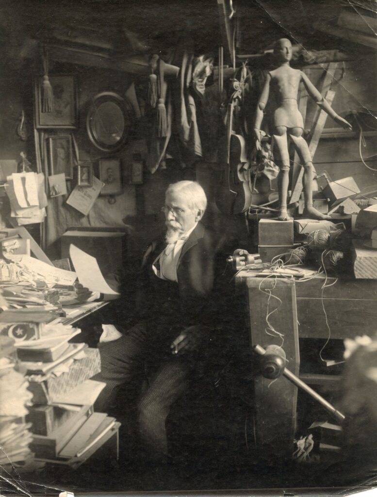 Edward Valentine reading in his studio surrounded by tools, papers, and a workbench with an artist’s mannequin sitting on top.