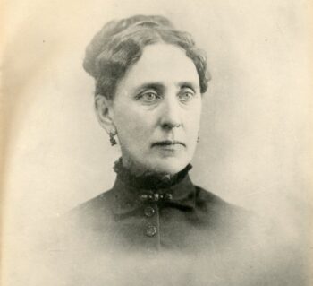 Portrait of a white woman with her hair up wearing earrings and a high neck black blouse.