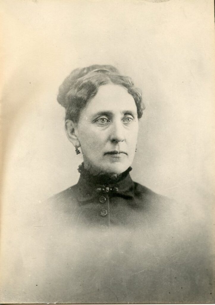 Portrait of a white woman with her hair up wearing earrings and a high neck black blouse.