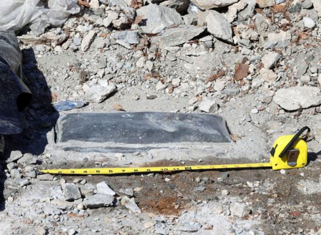Metal lid to a rectangular box visible in a pile of stone rubble with a measuring tape sitting in front of it.