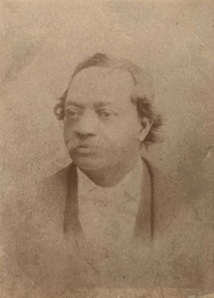 Portrait of a middle-aged Black man with a mustache wearing a white shirt and dark coat.