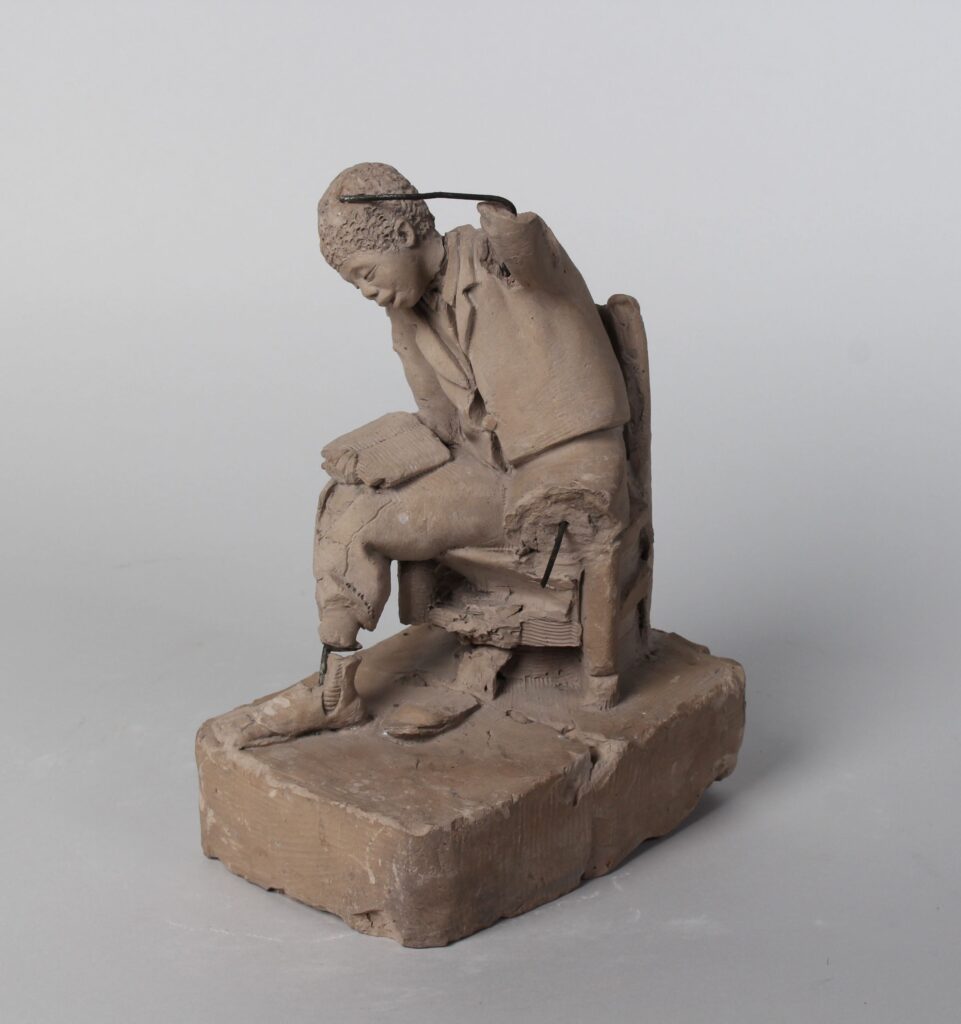 Clay model of a Black boy sitting in a chair leaning over an open book on his right knee with his left arm raised.