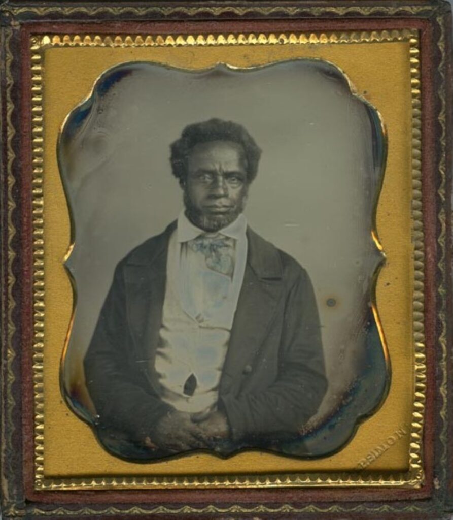 An unsmiling Black man from the waist up with a short beard, rumpled light-colored waistcoat, and dark wrinkled coat.