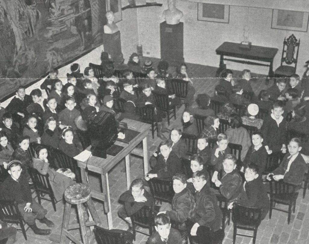 A class of school children sit in chairs looking back over their shoulders at the camera while a projector points forward toward walls with artwork covered by a pull-down screen.