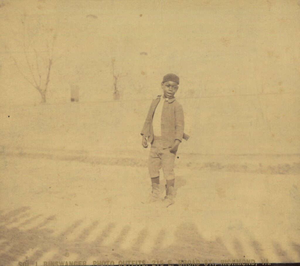 Sepia-toned image of a young Black boy standing in a yard wearing a knit cap, jacket, jacket, short pants, and boots.
