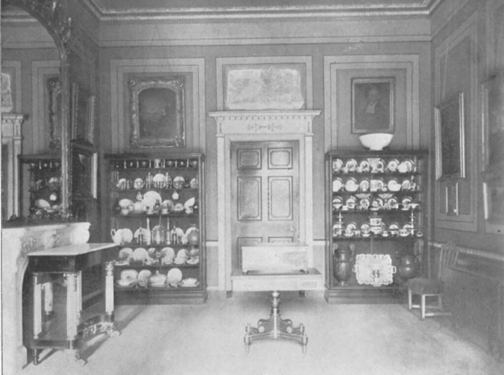 A view of the early Valentine Museum in the Virginia Room with cases full of china and glass, furniture in the room, and paintings on the walls.