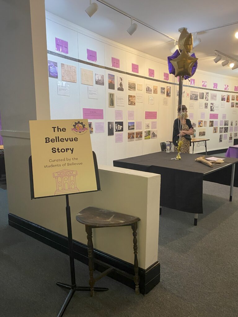 The Bellevue Story: Curated by the students at Bellevue. Shows a wall with a timeline on it. Also some purple and gold balloons.