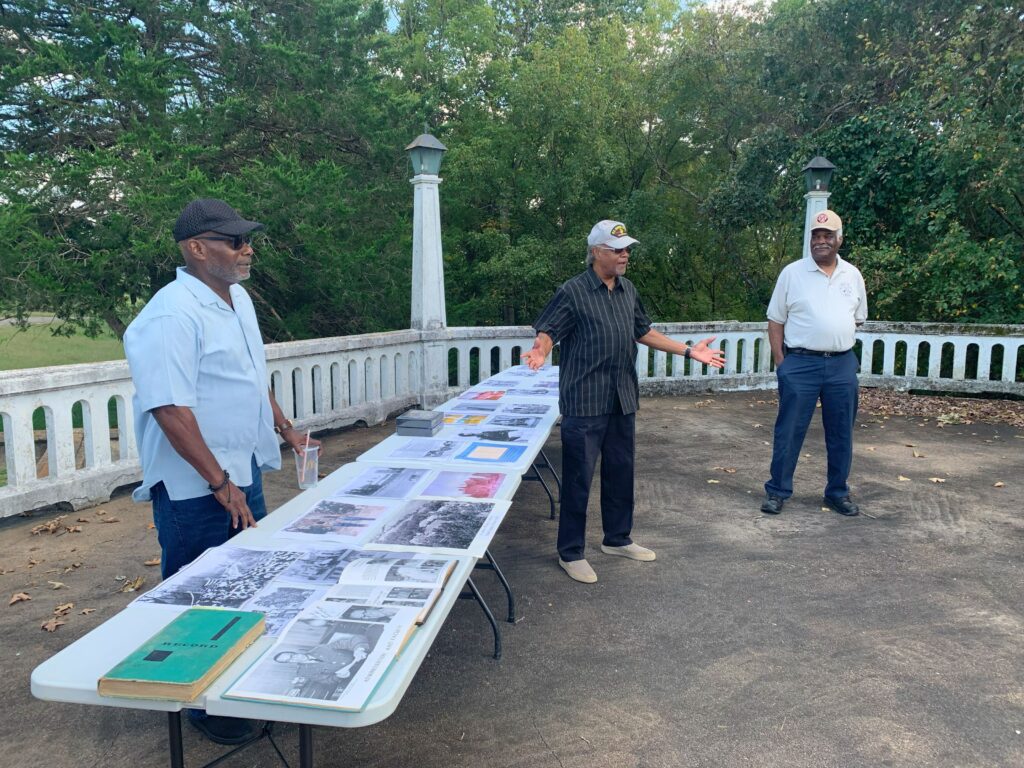 Three Black men all wearing baseball hats stand outside on a patio near three long tables full of books and photographs. One man is speaking.