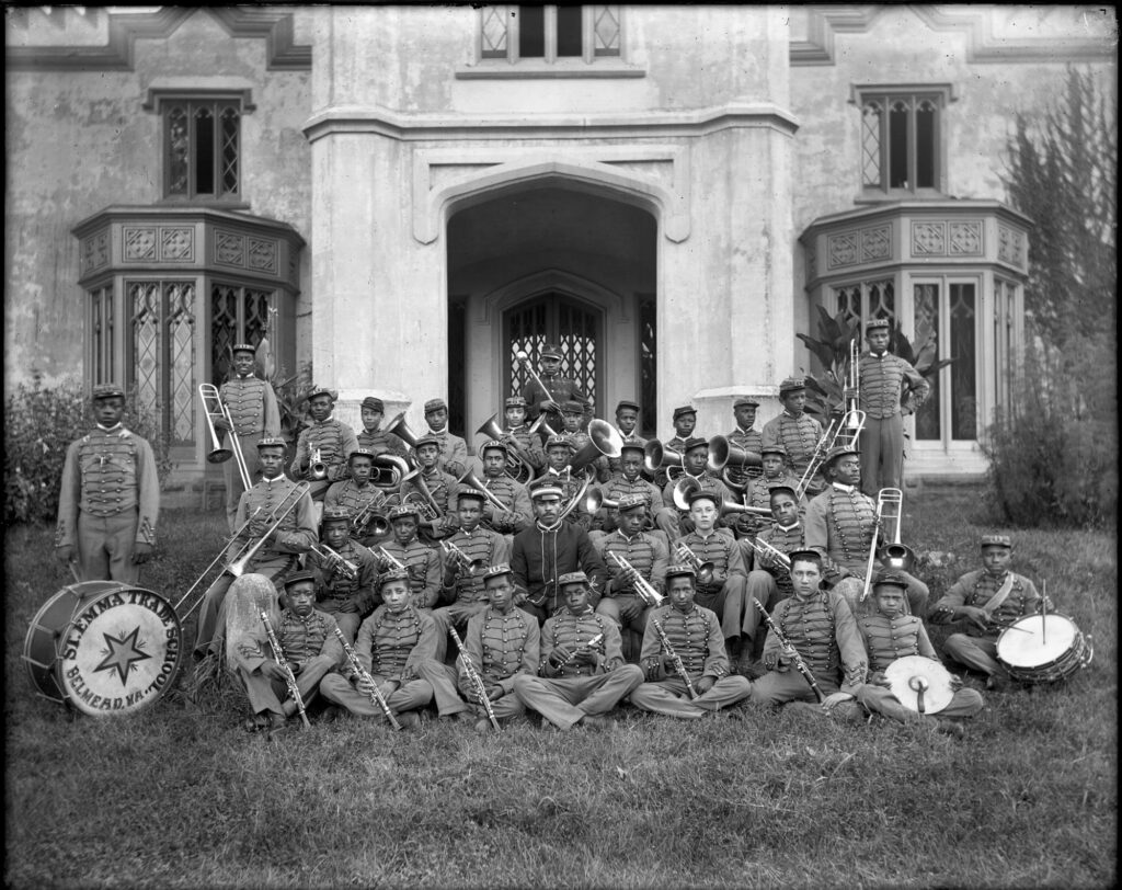 The marching band of St. Emma Industrial & Agricultural Institute at Belmead Plantation, Powhatan County near Richmond; image of a group of young Black men in band uniforms posing in front of the main school (house) building; the students hold instruments; band leader, in dark uniform, sits in center of group; drum on left inscribed, "St. Emma Trade School, Belmead, Va."