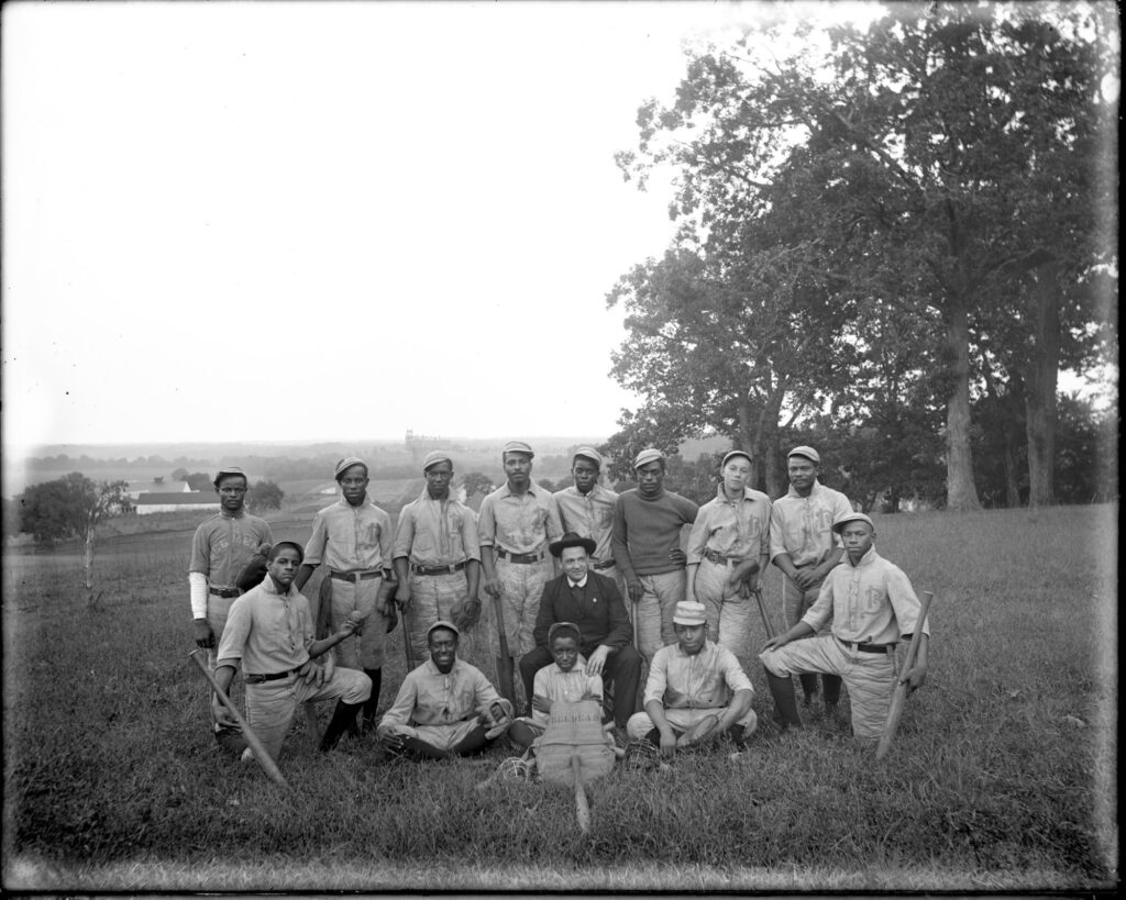 The school baseball team of St. Emma Industrial & Agricultural Institute at Belmead Plantation, Powhatan County, near Richmond; image of thirteen young Black men wearing baseball uniforms posed outside on the grounds of the school; possible coach in dark suit and hat, sits on stool amongst them.