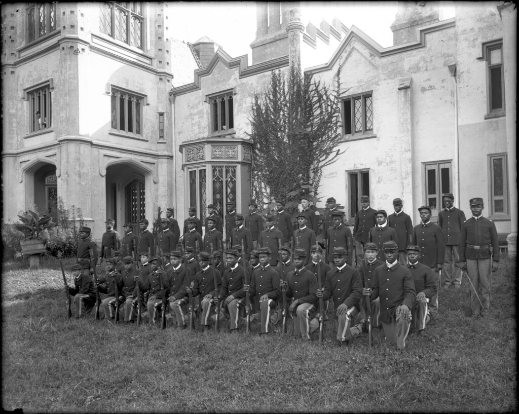 The cadet or rifle corps at the St. Emma Industrial & Agricultural Institute at Belmead Plantation, Powhatan County, near Richmond; image shows a group of young Black men in uniform posed with rifles in front of the Gothic Revival school building.