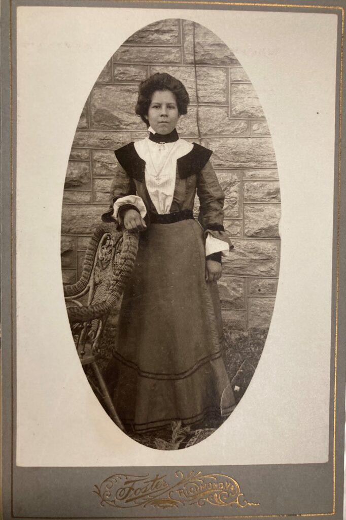 Young woman with dark hair pulled back with one arm resting on a wicker chair. She is wearing an Edwardian day dress with contrast standing neckline and modified pilgrim collar. The bodice has an inset front panel of striped, white material imitating a blouse and jacket. The skirt is in the fashionable bell shape of the time. In gold at the bottom, the picture says” Foster, Richmond, VA”