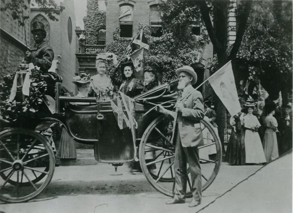 Three white ladies wearing dresses and hats sit in a carriage. A white bearded man wearing a suit and hat stands beside the carriage carrying a flag. The driver of the carriage is a Black man wearing a suit and hat. There is a wreath with a large bow next to the carriage driver.